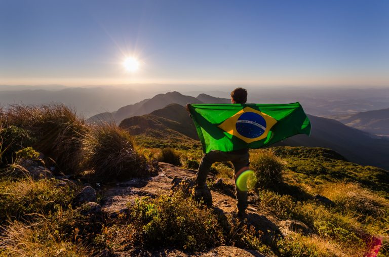 Young male holding a flag of Brazil and standing on top of a hill